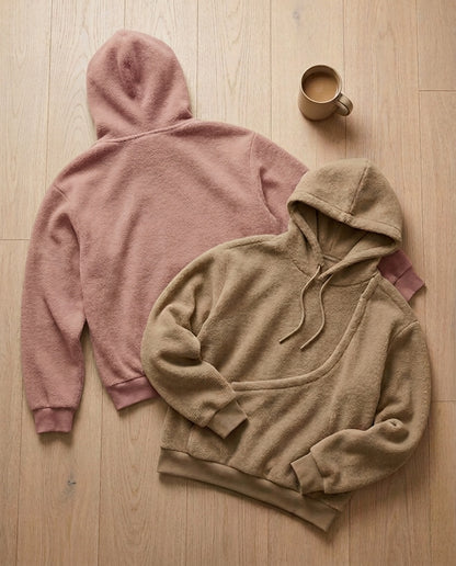 Two JOEY pouch hoodies laid on an oak floor, one khaki showing the diagonal chest pouch and one pink showing the plain back, with a mug of coffee
