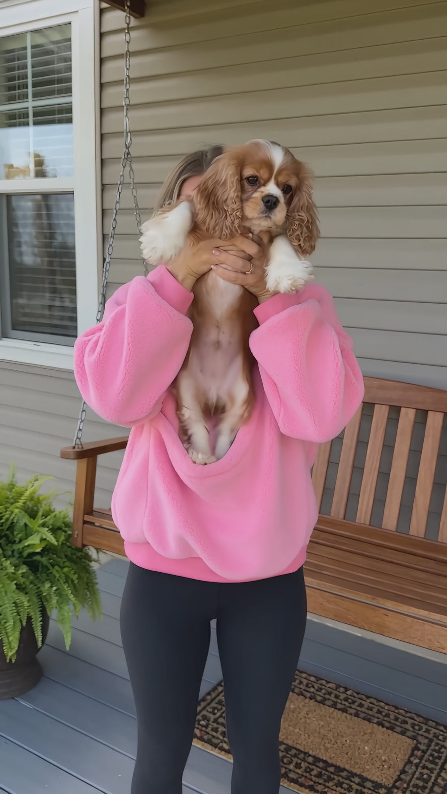 A woman on a porch lifts her small spaniel into the front pouch of a pink JOEY hoodie.